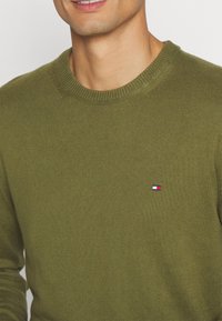 Maslinasto zeleni pleteni pulover s okruglim izrezom i rebrastim manžetama, s malim Tommy Hilfiger logotipom u crvenoj, bijeloj i plavoj boji na prsima.