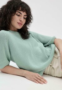 Pull en tricot vert clair avec une surface texturée, manches courtes et coupe décontractée, associé à un pantalon de teinte neutre.
