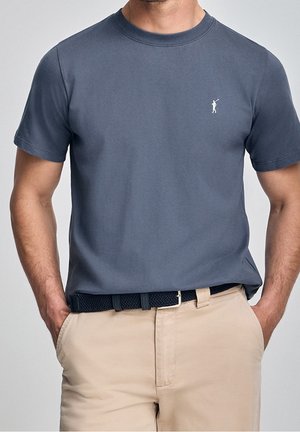 Kurzärmeliges, rundhalsausschnitt T-Shirt aus dunkelblauer Baumwolle mit kleinem weißen Logo. Kombiniert mit beigen Hosen und einem strukturierten schwarzen Gürtel.