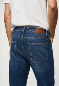 Jean en denim bleu foncé avec une finition texturée, doté de deux poches arrière et d'une étiquette en cuir marron sur la ceinture.