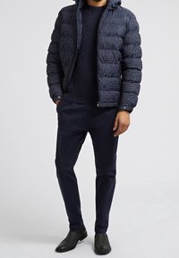 Marineblauwe gewatteerde pufferjack met capuchon en logopatronen, gedragen over een gestructureerde marineblauwe gebreide trui en donkere op maat gemaakte broeken, gecombineerd met zwarte laarzen.