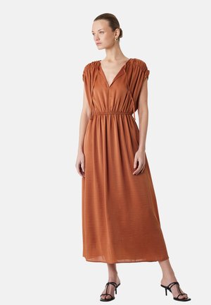 Kvinde står iført en rust-orange midi-kjole med rynkede kap-sleeves, V-hals, markeret talje og sorte sandaler med hæl og stropper.