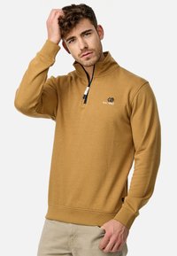 Sudadera color marrón claro con cuello con cremallera, que presenta un logo bordado y puños acanalados, hecha de un tejido suave.