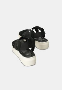 Crne zamšaste platforme s sandalama imaju prepletene trake i masivan bijeli potplat. Srednji potplat pruža visinu uz održavanje elegantnog dizajna.