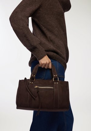 Borsa a tracolla in suede marrone con hardware dorato, doppi manici, tasca frontale con zip e forma rettangolare strutturata. Indossata con un maglione scuro e jeans blu.