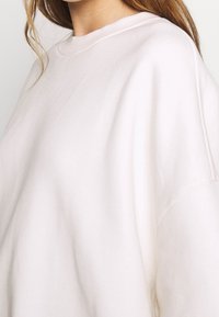 Sudadera rosa claro de gran tamaño hecha de tela suave, con cuello redondo y hombros caídos, con una superficie lisa y ligeramente texturizada.