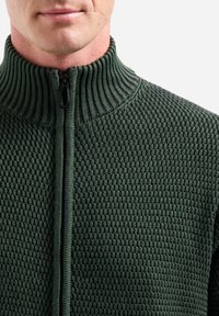 Donkergroene gebreide ritstrui met een hoge kraag en een gestructureerd patroon. Voorzien van een gladde ritssluiting en geribde accenten bij de kraag.