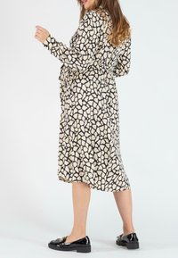 Kleid in Creme mit einem schwarzen abstrakten Muster, langen Ärmeln, Knopfverschluss und einem Gürtel. Modell kombiniert mit schwarzen Loafers.