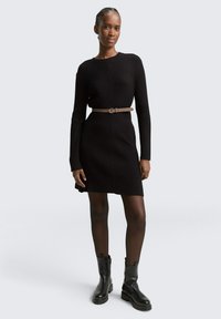 Schwarzes geripptes Strickkleid mit langen Ärmeln, in der Taille mit einem hellbraunen Gürtel gerafft. Getragen mit schwarzen Stiefeletten und durchsichtigen Strumpfhosen.