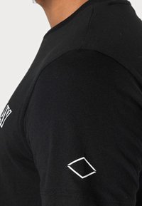 Camiseta negra de manga corta en suave algodón, con un logo geométrico blanco en la manga izquierda. Corte ajustado y diseño sencillo de cuello redondo.
