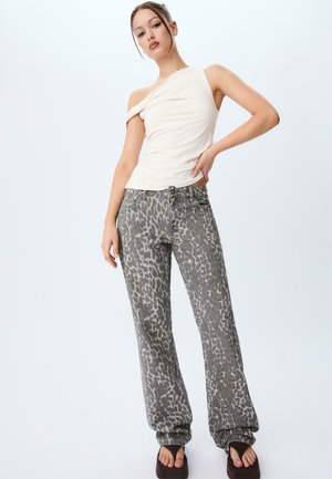 Beige top met één schouder, gecombineerd met grijze spijkerbroek met luipaardprint, met een losse pasvorm en lichte stretch. Model staat met handen op de heupen.
