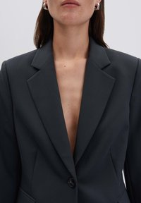 Blazer grigio scuro con spalle strutturate, rever a punta e profondo scollatura a V. Chiusura con un bottone, vestibilità sagomata, texture liscia.