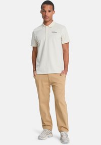 Crème polo shirt met korte mouwen en een drieknoopsluiting, gecombineerd met tan broek en lichtgrijze sneakers, benadrukt casual stijl.