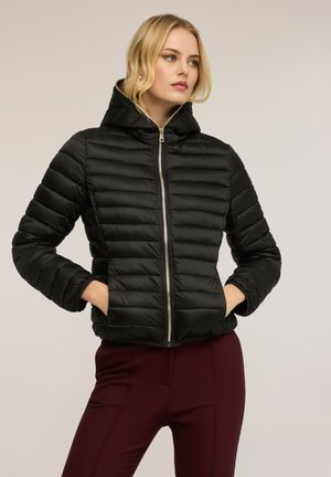 Vaaleahiuksinen nainen, jolla on musta tikattu toppatakki hupulla ja viininpunaiset housut, kädet taskuissa, katsoo sivulle.