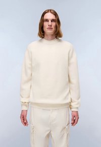 Suéter de punto color crema con cuello redondo, puños y dobladillo acanalados. El corte holgado se combina con pantalones a juego de color claro.