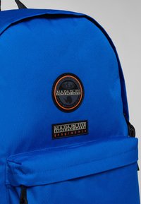Zaino blu realizzato in nylon resistente, con patch rotondo del logo e etichetta rettangolare, tasca frontale con zip e dettagli neri.
