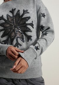 Pull gris avec un motif flocon de neige en sequins sur le devant et des accents en forme d'étoile sur les manches. Poignets et ourlet côtelés. Tissu doux et texturé.