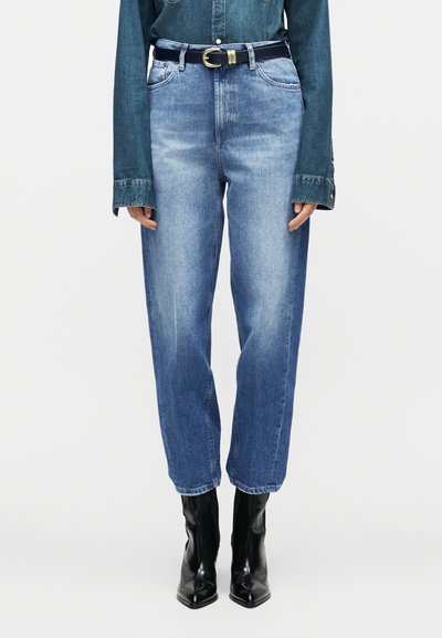 Άτομο που φοράει ψηλόμεσα μπλε τζιν με μαύρη ζώνη και μαύρα μποτάκια με μυτερή μύτη, στέκεται μπροστά σε απλό φόντο.