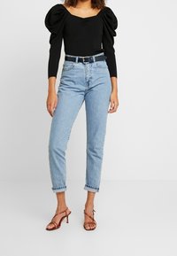 Zwarte top met pofmouwen, hooggewaiste lichtblauwe jeans en rode sandalen met hakken. Eenvoudig ontwerp, met opgerolde mouwen en een ceintuur in de taille.