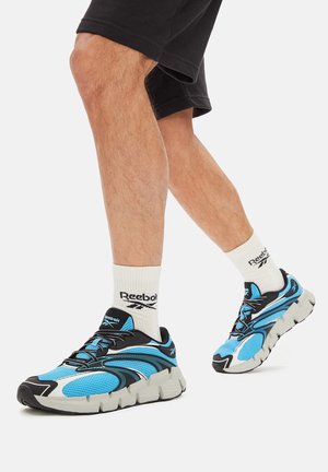 Niebieskie siateczkowe trampki z czarnymi dodatkami i teksturowaną podeszwą, w połączeniu z białymi skarpetkami Reebok i czarnymi szortami.