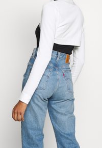 Gekürztes weißes Langarmoberteil, das über einem schwarzen Oberteil getragen wird; kombiniert mit hoch sitzenden hellblauen Jeans aus Denim, die hinten mit einem braunen Lederpatch versehen sind.