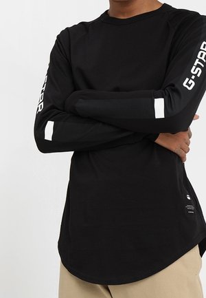 Person iført en sort langærmet skjorte med teksten "G-STAR" på ærmerne og beige bukser, arme krydset over brystet.