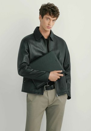 Młody mężczyzna z kręconymi włosami, ubrany w czarną skórzaną kurtkę i beżowe spodnie, trzymający czarny pokrowiec na laptopa na jednolitym tle.