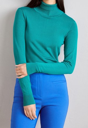 Osoba v modrozeleném roláku a jasně modrých kalhotách, s překříženýma rukama přes hruď na jednobarevném světlém pozadí.