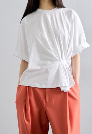 Kobieta ubrana w białą bluzkę z krótkim rękawem i marszczeniami po bokach oraz rdzawobrązowe, plisowane spodnie z wysokim stanem, z rękami w kieszeniach.