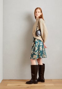 Beige strikkegenser, teal blomsterkjole, brune støvler til knærne. Utseendet har en teksturert genser og et glatt stoffer skjørt med et blomsterdesign.