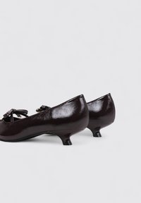 Zapatos de charol marrón oscuro con un pequeño tacón, que cuentan con un acento de lazo en la parte frontal. Diseño elegante con un acabado brillante.