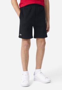 Shorts en coton noir avec une taille élastique et des poches latérales, ornés d'un logo Lacoste blanc sur la jambe gauche.