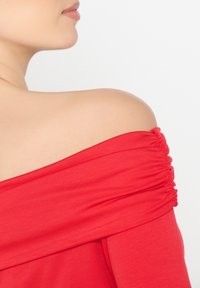 Top rosso senza spalle con dettaglio di tessuto arricciato e texture liscia, caratterizzato da una vestibilità ampia che si adagia attorno al busto.