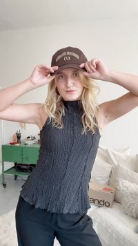 Schwarzes, strukturiertes, ärmelloses Oberteil mit hohem Kragen, kombiniert mit einer braunen Mütze mit weißen Schriftzug. Einfacher Hintergrund mit weichen Kissen.