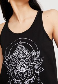Femme portant un haut sans manches noir avec un design floral mandala blanc détaillé, mettant l'accent sur le cou, l'épaule et le haut de la poitrine.