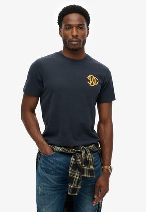 Mand iført en mørkeblå T-shirt med gul logo, blå jeans og en ternet skjorte bundet om livet, stående med den ene hånd i lommen.