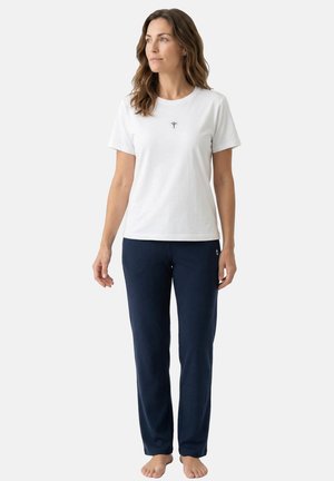 Kvinde står barfodet iført en hvid kortærmet skjorte med lille logo og marineblå bukser med lige ben mod en ensfarvet baggrund.