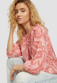 Blumenbluse mit Puffärmeln, die ein rosa-weißes Muster auf leichtem Stoff zeigt. Modell sitzt mit Jeans und präsentiert einen lässigen Stil.