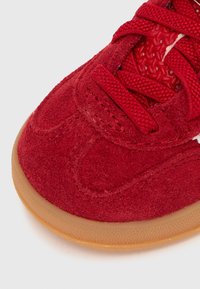 Červená semišová teniska s texturovaným svrškem, kulatou špičkou a shodnými tkaničkami. Má podrážku z gumové pryže a vzorovanou vnitřní podšívku.