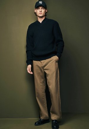 Navy knitted sweater, tan wide-leg pants, and black loafers. A black cap completes the look. Simple, casual ensemble against a muted background.
