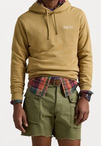 Felpa con cappuccio gialla con ricamo "Polo RL", indossata sopra una camicia di flanella a quadri. Abbinate a pantaloni cargo verdi e un orologio.