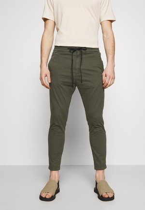 Man draagt olijfgroene broek met trekkoord, beige kortemouws overhemd en open beige en zwarte sandalen, staand tegen een effen witte muur.