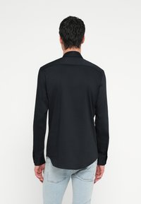 Fekete hosszú ujjú ing sima textúrával, gombos mandzsettával és ívelt aljjal, hátulról fehér háttér előtt bemutatva.