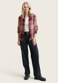 Chemise à carreaux rouge et rose sur un haut blanc, associée à un pantalon large bleu marine et des chaussures noires épaisses.
