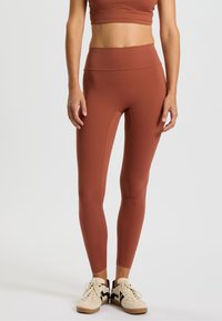 Roestkleurige leggings gemaakt van rekbaar materiaal, met een hoge tailleband en een gladde textuur, gecombineerd met beige en zwarte sneakers.