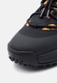 Μαύρο αθλητικό παπούτσι με υφή στη λαβή από καουτσούκ, διαθέτον τας αεροδυναμικό σχεδιασμό και ελαστικά κορδόνια. Οι λεπτομέρειες περιλαμβάνουν φωτεινό πορτοκαλί μοτίβο.