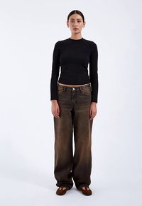 Top negro ajustado de manga larga. Vaqueros marrones descoloridos y holgados con corte recto. Zapatos marrones tipo mocasín. Fondo simple.