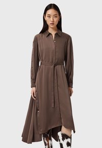 Abito camicia in raso marrone con maniche lunghe, chiusura frontale con bottoni e cintura in vita. Presenta un orlo asimmetrico più lungo dietro e spaccature laterali.