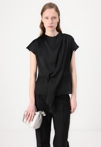 Top nero con dettaglio increspato sul davanti, maniche corte, abbinato a pantaloni neri su misura. Tiene una piccola borsa a tracolla in satiné argento.