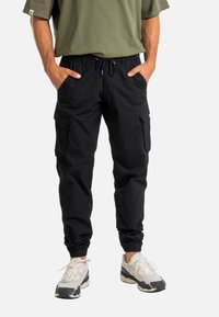 Mann in schwarzen Cargo-Jogginghosen mit Seitentaschen, elastischen Bündchen, weißen und schwarzen Turnschuhen sowie einem olivgrünen Kurzarmhemd, die Hände in den Taschen.